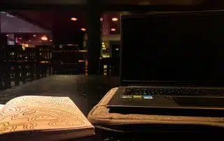 A dimly lit room with a closed laptop on a padded sleeve and an open notebook with a swirly design sitting on a dark wooden table—my favourite study spot. In the background, an empty bar area with high chairs can be seen. This serene setting is perfect for my February Photo Contest entry.