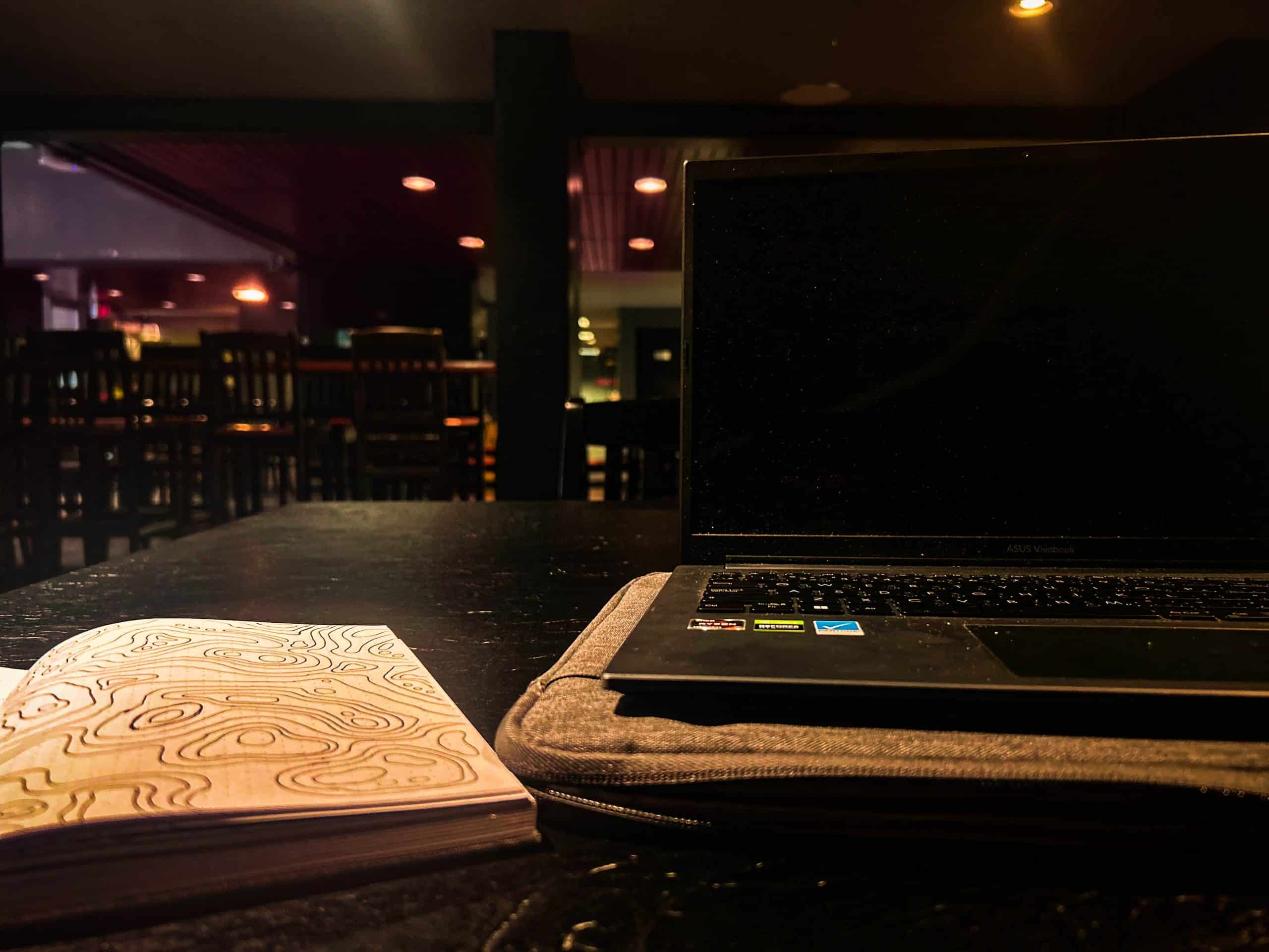 A dimly lit room with a closed laptop on a padded sleeve and an open notebook with a swirly design sitting on a dark wooden table—my favourite study spot. In the background, an empty bar area with high chairs can be seen. This serene setting is perfect for my February Photo Contest entry. A dimly lit room with a closed laptop on a padded sleeve and an open notebook with a swirly design sitting on a dark wooden table—my favourite study spot. In the background, an empty bar area with high chairs can be seen. This serene setting is perfect for my February Photo Contest entry.