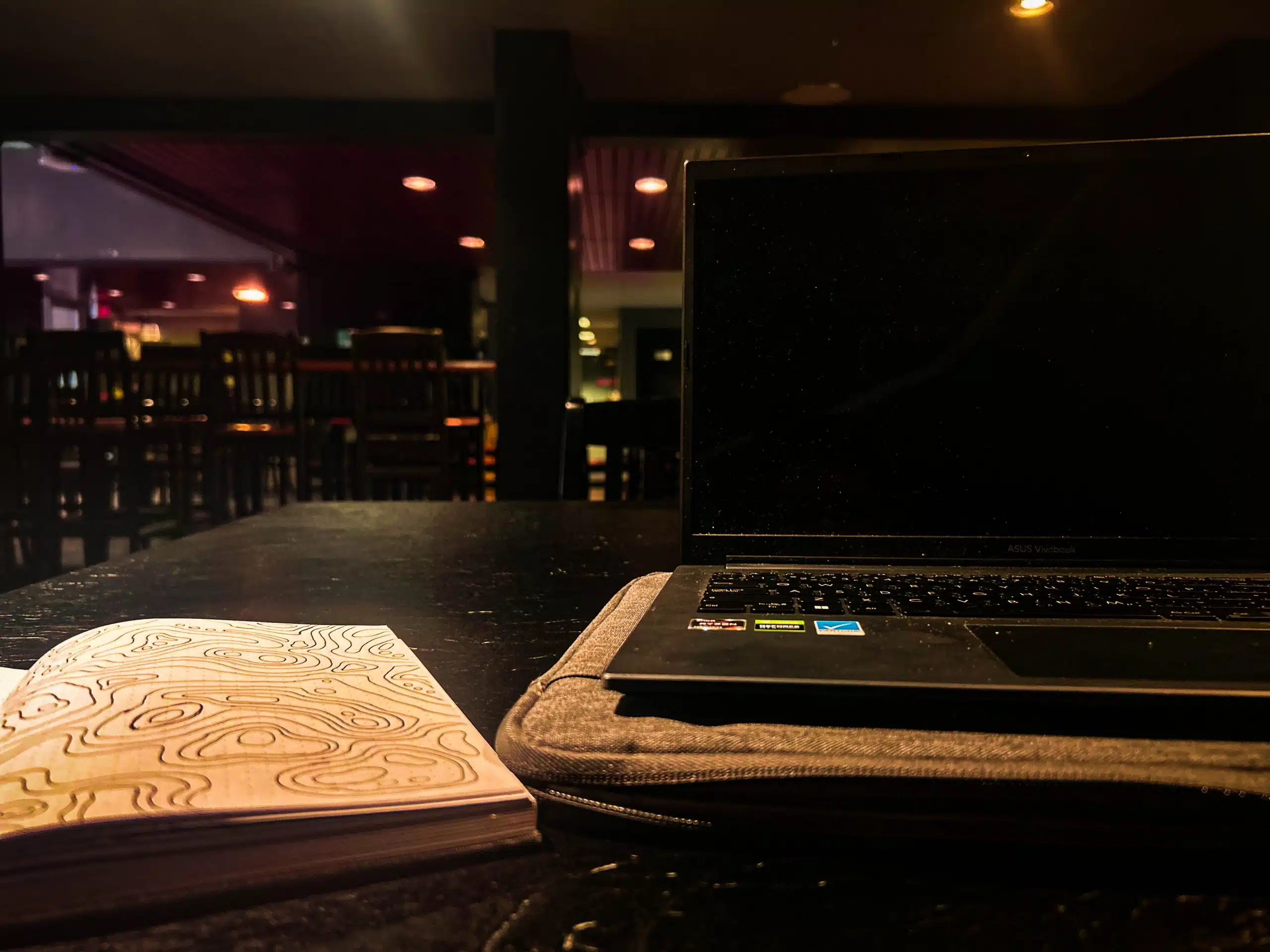 A dimly lit room with a closed laptop on a padded sleeve and an open notebook with a swirly design sitting on a dark wooden table—my favourite study spot. In the background, an empty bar area with high chairs can be seen. This serene setting is perfect for my February Photo Contest entry.