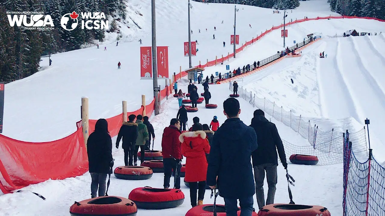 People in winter clothing walk up a snow-covered hill while pulling inner tubes at a tubing park. Many tubes are red and black, and there are safety net barriers along the path. The hill ahead has multiple tubing lanes and is surrounded by trees. Logos appear in the top left.