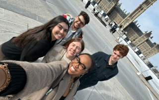 A group of five smiling individuals taking a selfie outside on a paved area with historic, gothic-style buildings in the background. Some of the buildings have tall, pointed towers. The sky is cloudy, and the people appear happy and relaxed—a perfect snapshot from Lobby Week 2024 by UCRU, advocating for change.