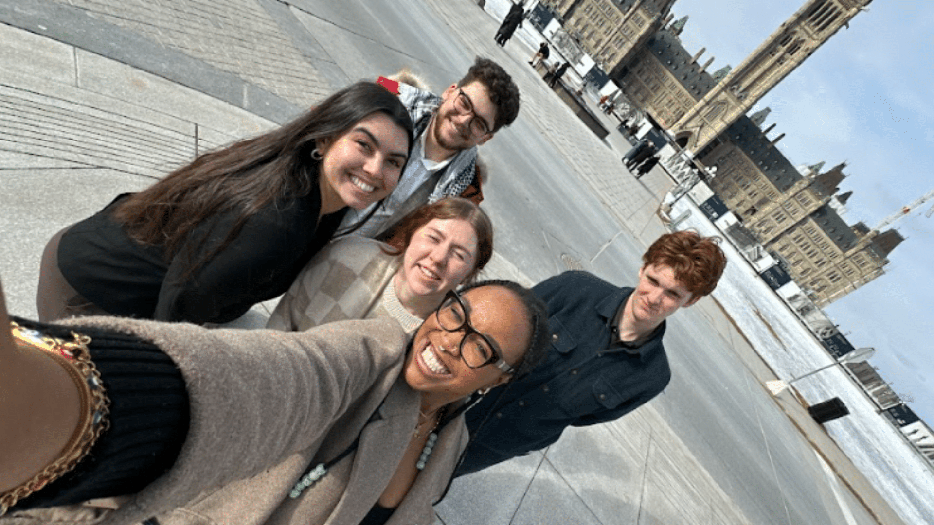 A group of five smiling individuals taking a selfie outside on a paved area with historic, gothic-style buildings in the background. Some of the buildings have tall, pointed towers. The sky is cloudy, and the people appear happy and relaxed—a perfect snapshot from Lobby Week 2024 by UCRU, advocating for change.