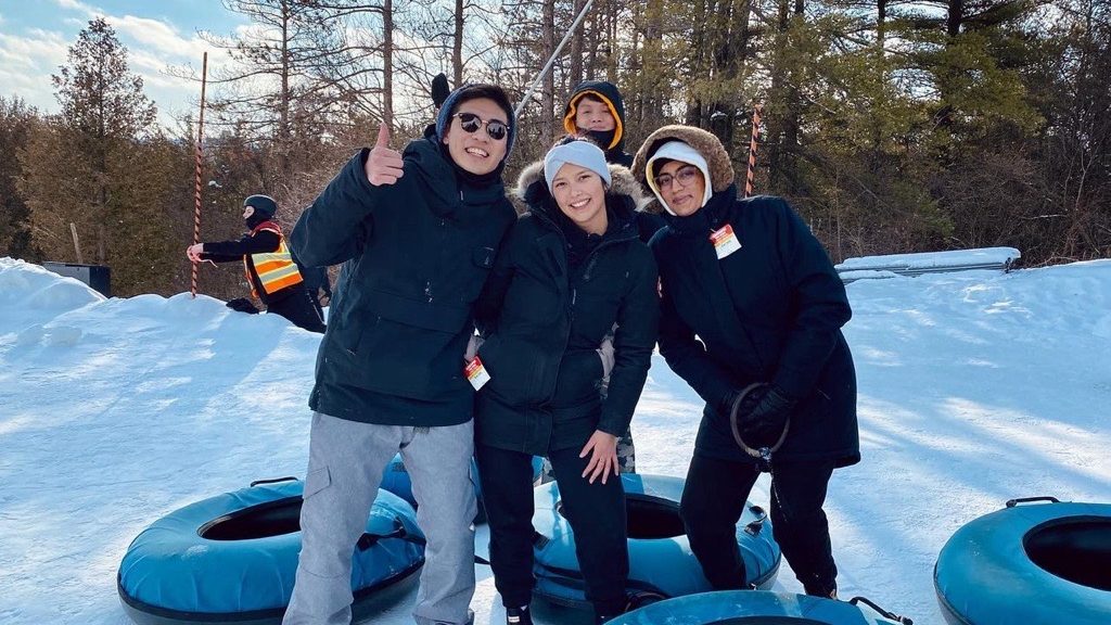 Four people in winter clothing stand together on a snowy slope with blue snow tubes. One person gives a thumbs-up, and another kneels in the center. Trees are visible in the background, and a person in an orange vest can be seen on the left side of the image.