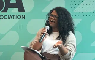 A woman with curly dark hair, wearing glasses, a beige cardigan, and holding a microphone, is speaking passionately. Her other hand is gesturing expressively about anti-racism. She is sitting against a green backdrop with partial text visible, likely engaging in important conversations on the topic.