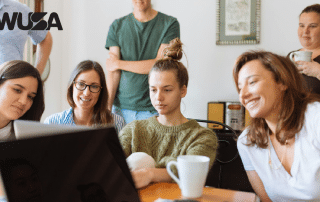 A group of six people are gathered around a table, looking at a laptop screen and smiling. Some hold coffee cups. They appear engaged and happy. There's a WUSA logo in the top-left corner of the image.