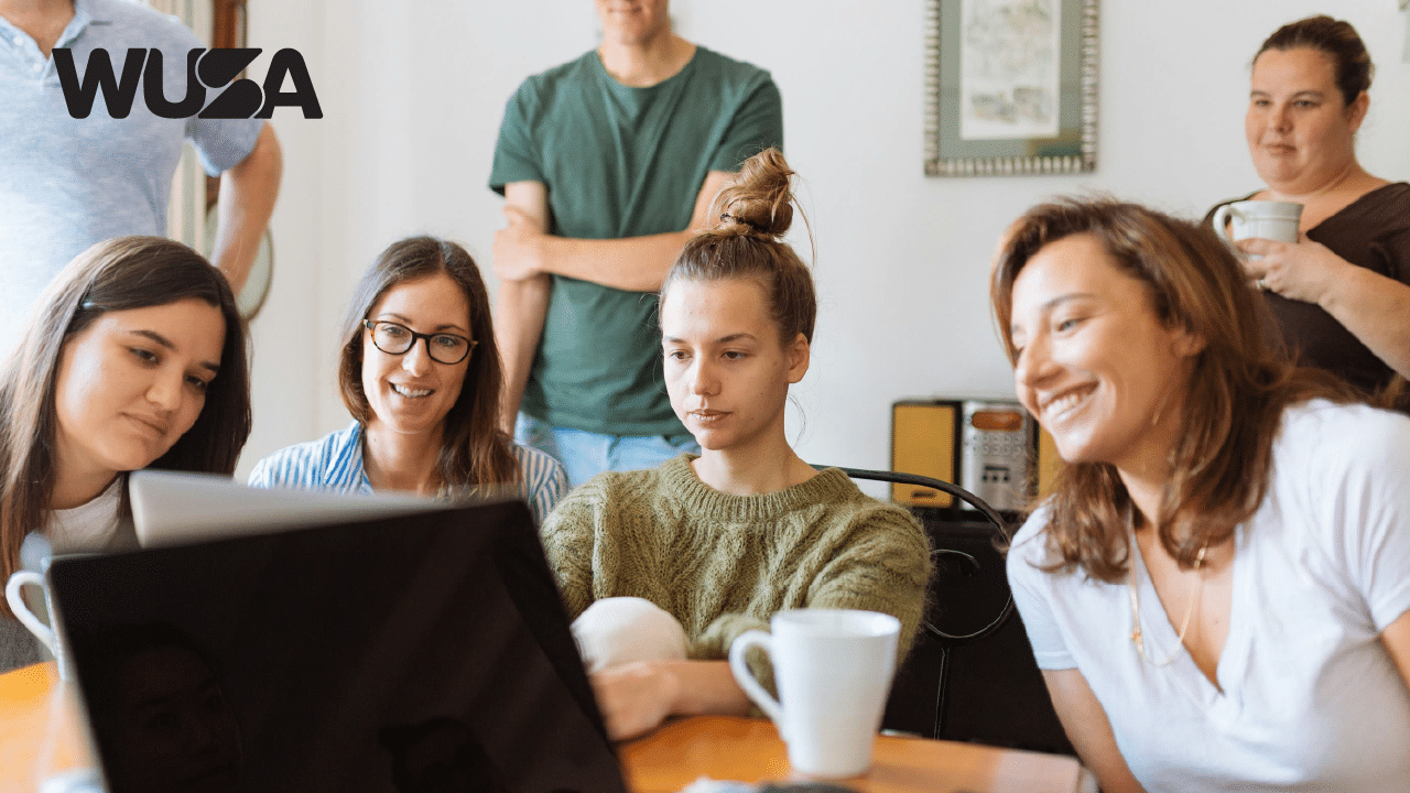 A group of six people are gathered around a table, looking at a laptop screen and smiling. Some hold coffee cups. They appear engaged and happy. There's a WUSA logo in the top-left corner of the image.
