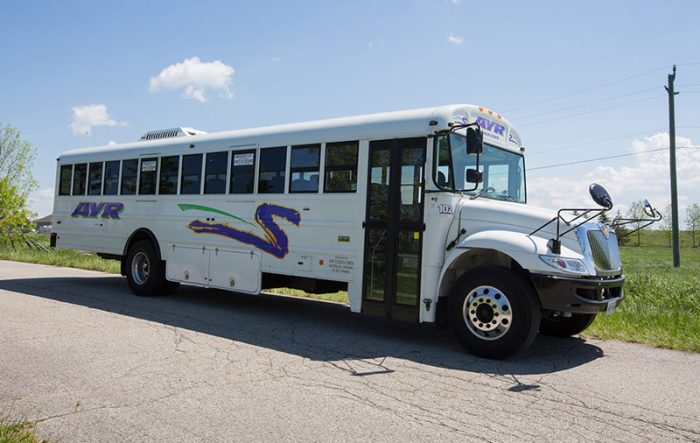 A white bus featuring the AVR logo and a vibrant side design is parked on a road under the clear blue sky during the Spring Term. Ideal for holders of the Stratford Bus Pass - Spring Term Sale, the bus includes side windows, a side door, and a front stop sign arm. Grass and power lines can be seen in the background near Stratford.