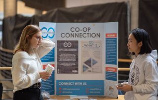 Two individuals are conversing in front of a display board labeled "CO-OP CONNECTION." The board behind them has information and pamphlets about WUSA programs, volunteer opportunities, and contact information. One person is holding a coffee cup while the other appears to be distributing flyers.