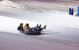 Two people, dressed in winter clothing, are riding inflatable tubes down a snowy slope at night during the Winter 2024 Term. The smooth, white snow stretches out around them, with piles of snow in the background and some blue objects visible near the top-right corner.