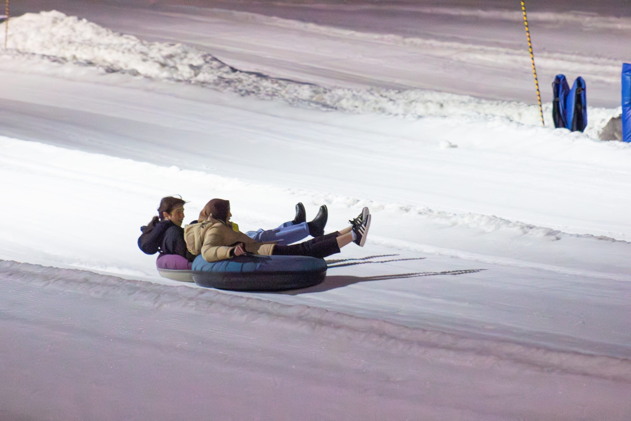 Two people, dressed in winter clothing, are riding inflatable tubes down a snowy slope at night during the Winter 2024 Term. The smooth, white snow stretches out around them, with piles of snow in the background and some blue objects visible near the top-right corner.