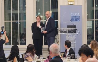 Two individuals stand on a stage in front of an "Ontario Undergraduate Student Alliance" banner. The individual on the left holds a plaque for the 2023 Teaching Excellence Award, while the other is shaking hands and smiling. Several people seated at tables in the foreground take photos and applaud.