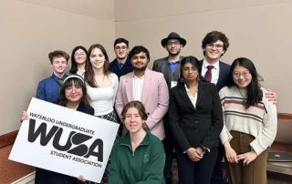 A group of thirteen students posing indoors. One person in front holds a sign that reads "Waterloo Undergraduate Student Association WUSA." The group, dressed in a mix of formal and casual attire, stands against a plain beige background, ready to represent at the 59th General Assembly.