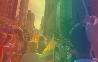 A crowd of people gathered on a city street during the Toronto Pride March. Many in the crowd hold rainbow flags and signs. Tall buildings line both sides of the street. The image is overlaid with a rainbow filter, enhancing the celebratory glow and atmosphere.