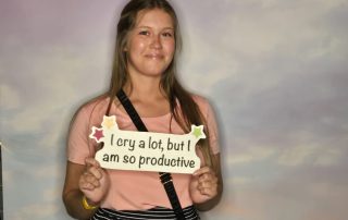 A young woman stands against a cloudy backdrop, smiling and holding a sign that reads, "I cry a lot, but I am so productive." She is wearing a light pink shirt, a black-and-white striped skirt, and has a black crossbody bag. This candid shot could be one of the standout entries for the May 2024 Photo Contest.