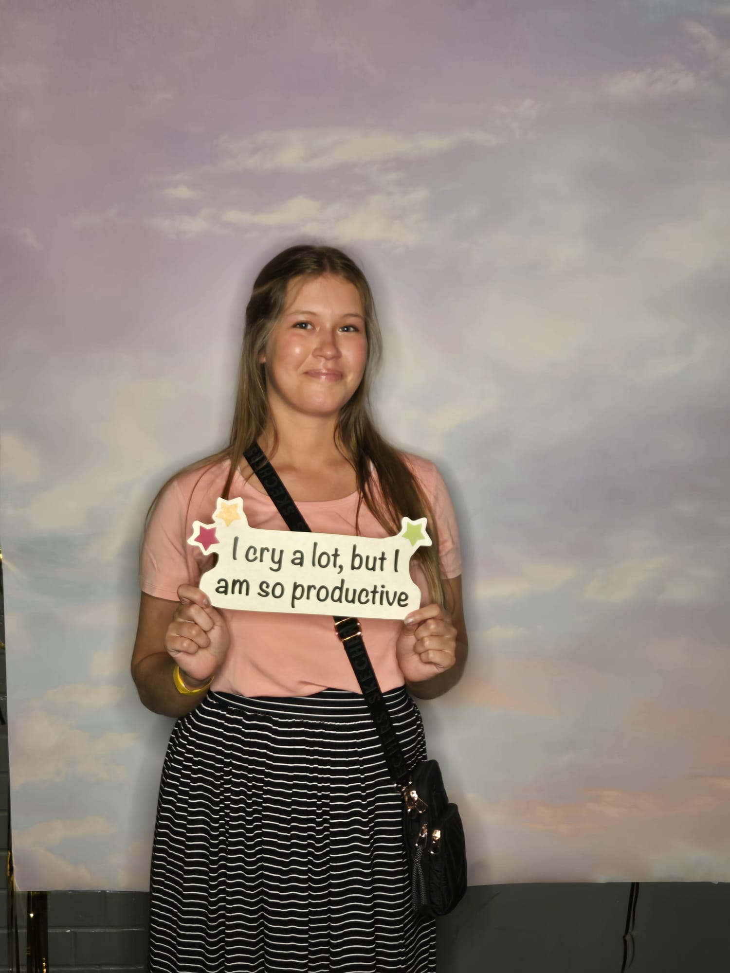 A young woman stands against a cloudy backdrop, smiling and holding a sign that reads, "I cry a lot, but I am so productive." She is wearing a light pink shirt, a black-and-white striped skirt, and has a black crossbody bag. This candid shot could be one of the standout entries for the May 2024 Photo Contest.