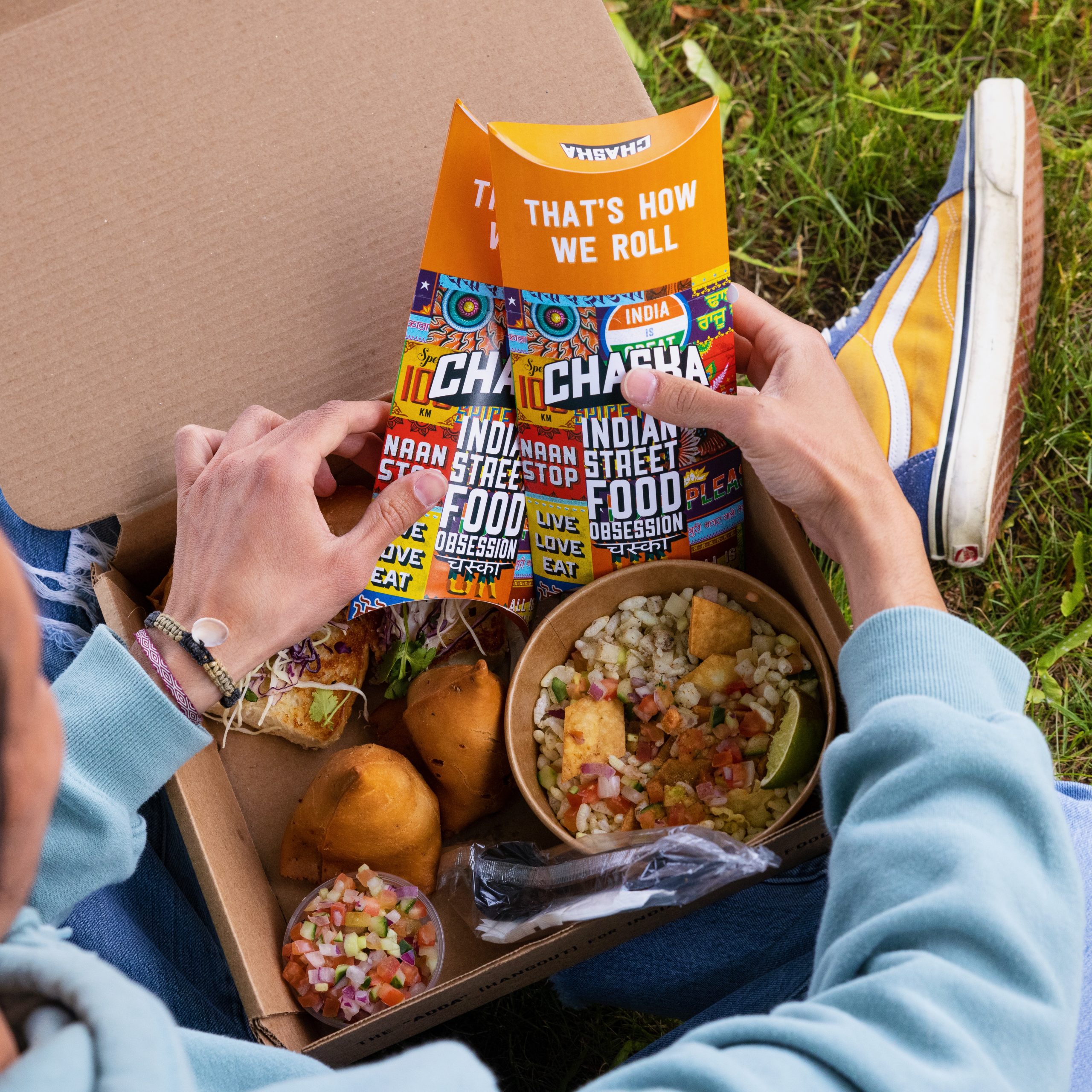 A person is sitting on the grass, holding two colorful, folded menus from Chacha Feeka, an Indian street food restaurant. In front of them is a cardboard tray containing samosas, salad, snacks, and cutlery. They are wearing a light blue sweatshirt and yellow sneakers.