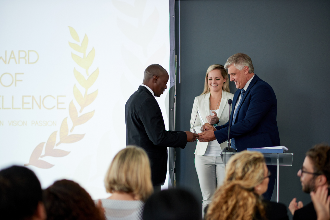 A man in a black suit is being handed an award by a man in a blue suit and a woman in a white suit on stage. An audience is seated in front of them, watching intently. The background has a screen displaying the text "AWARD OF EXCELLENCE" at the OUSA Teaching Awards.