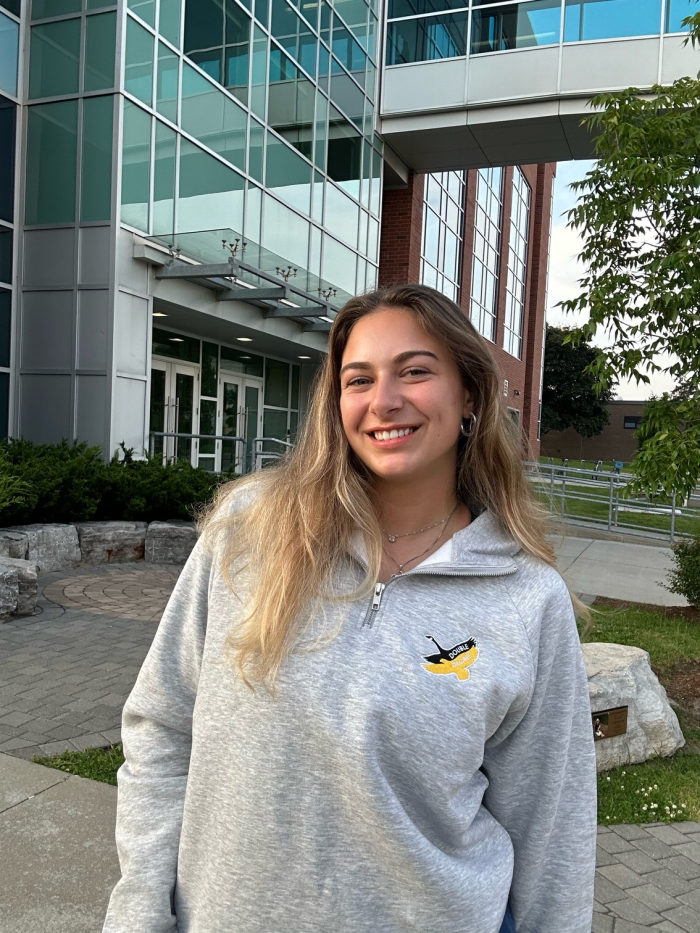 A young person with long blonde hair is smiling at the camera, standing outside a building with large glass windows. They are wearing a light gray DDC Quarter Zip sweater featuring a small emblem. The surroundings include greenery, a pathway, and parts of other buildings.