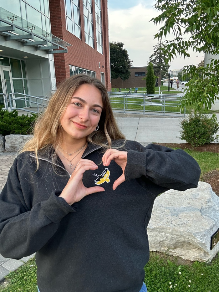 A young woman with long hair stands outside a modern brick building, making a heart shape with her hands. She is wearing one of the DDC Quarter Zips featuring a small yellow and white graphic and smiling at the camera. Trees, sidewalks, and green grass are visible around her.