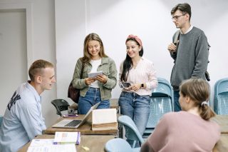 A group of young adults are gathered around a table in a classroom setting. Two women and a man are standing, smiling and engaged in conversation, while two people are seated at the table with books and a laptop in front of them. The room has white walls and blue chairs, likely planning for the May 2024 board meeting recap.