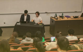 A lecture hall with rows of students seated, facing two people at the front who are sitting at a table with laptops. One is dressed in a suit, and the other in a casual shirt. A whiteboard and various equipment are visible in the background, suggesting an upcoming discussion on decision-making processes.