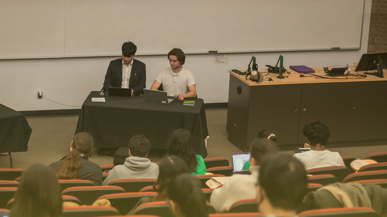 A lecture hall with rows of students seated, facing two people at the front who are sitting at a table with laptops. One is dressed in a suit, and the other in a casual shirt. A whiteboard and various equipment are visible in the background, suggesting an upcoming discussion on decision-making processes.