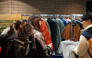 Several people are shopping for clothes at a sale or thrift event. They are gathered around clothing racks filled with various garments. The setting appears to be indoors, with a sign in the background and brick walls. Some individuals are wearing backpacks.