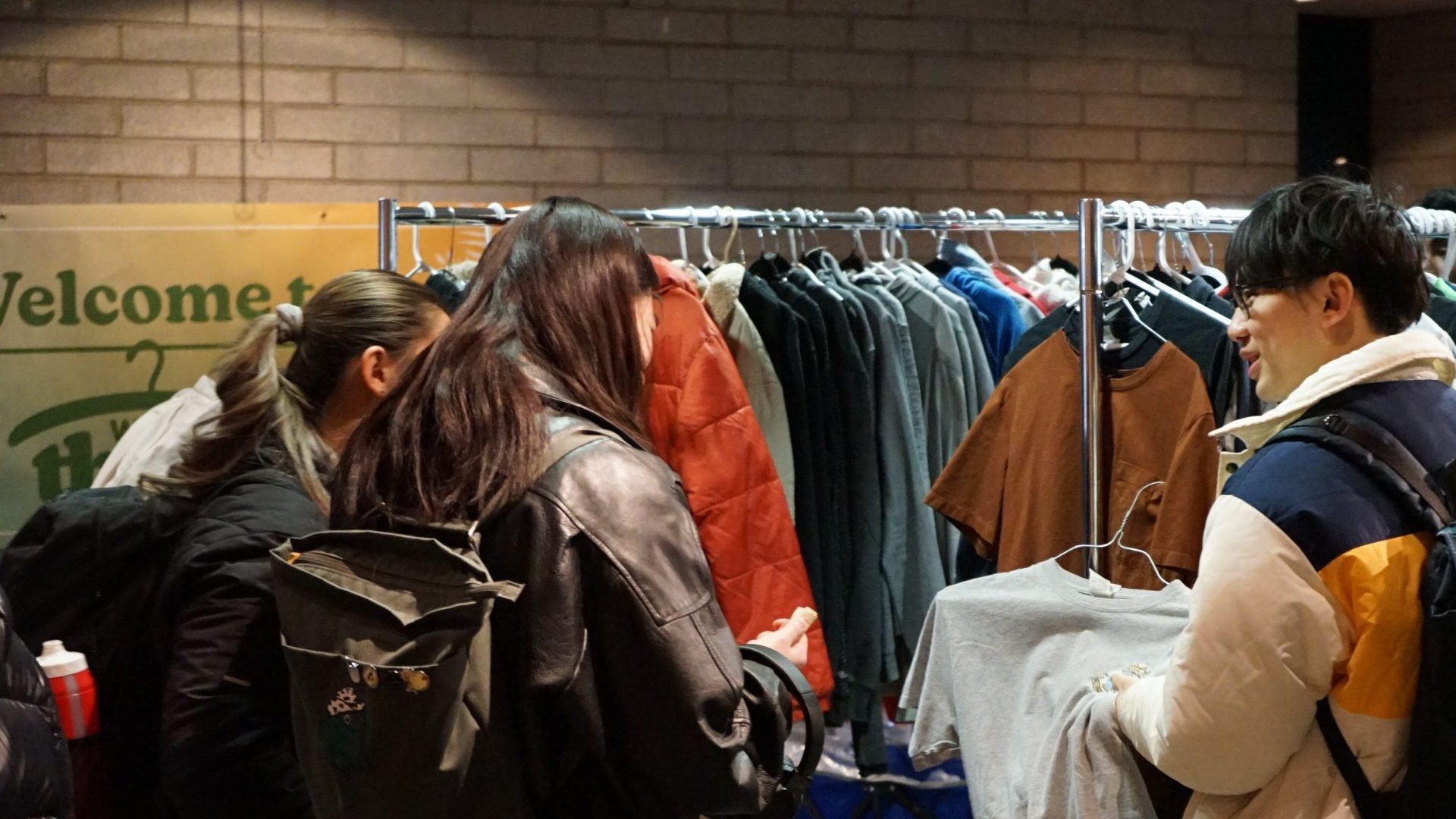 Several people are shopping for clothes at a sale or thrift event. They are gathered around clothing racks filled with various garments. The setting appears to be indoors, with a sign in the background and brick walls. Some individuals are wearing backpacks.