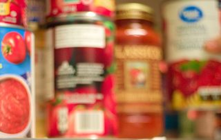 A blurry image of various canned goods on a shelf. The cans, seemingly from different brands and types including tomatoes and sauce products, have indistinct labels due to lack of focus. The image evokes a general impression of a pantry shelf, reminiscent of those provided by local food support services.