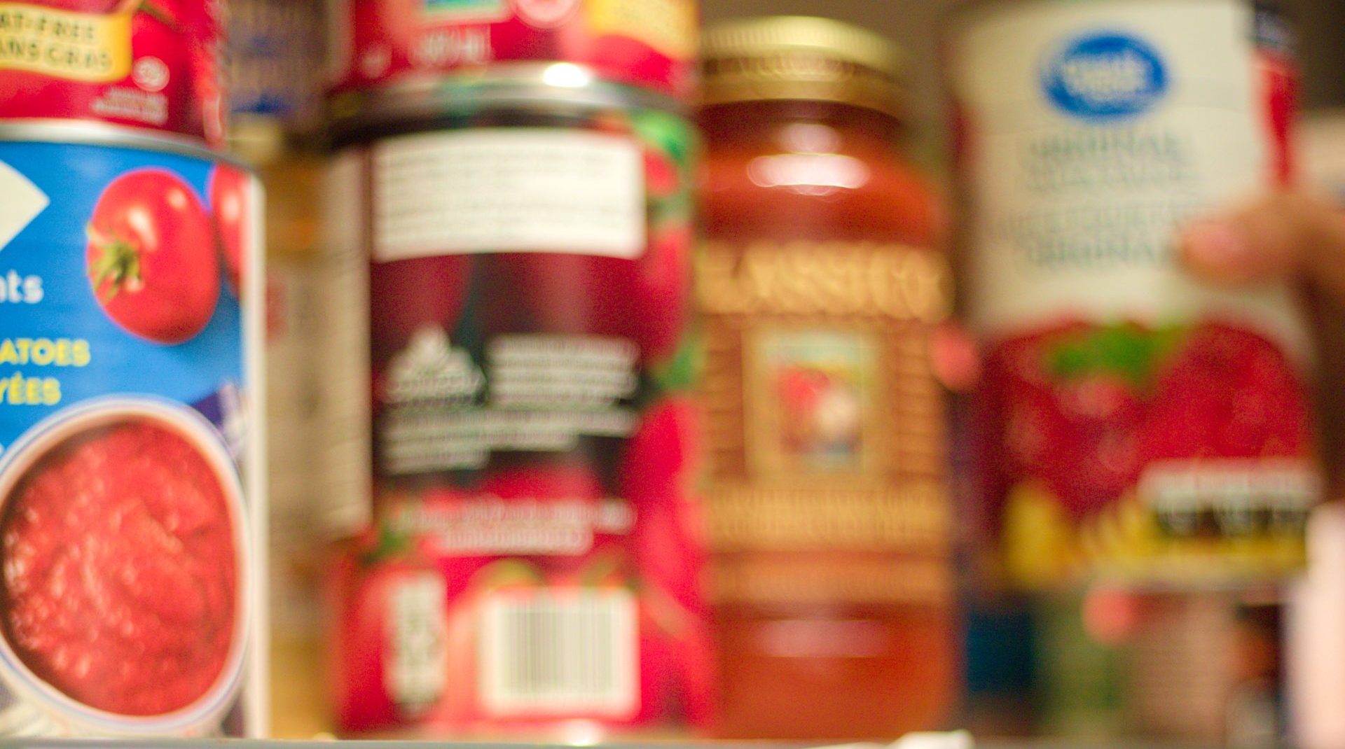 A blurry image of various canned goods on a shelf. The cans, seemingly from different brands and types including tomatoes and sauce products, have indistinct labels due to lack of focus. The image evokes a general impression of a pantry shelf, reminiscent of those provided by local food support services.