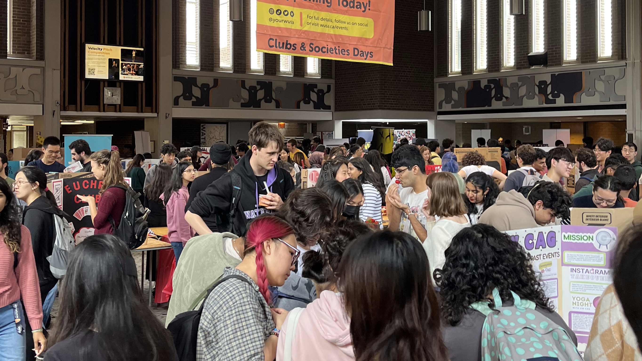 A crowded indoor event with numerous people walking around and engaging with various booths and posters. A large banner above the crowd reads, "Clubs & Societies Fair." The scene is bustling with activity as attendees interact and explore the displays.