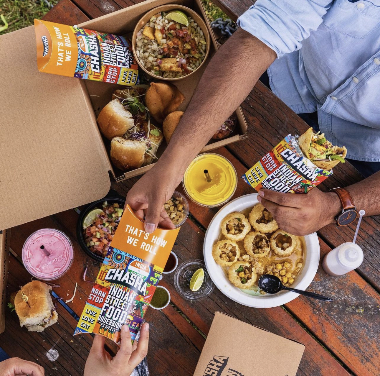 Two people sharing a meal at a picnic table, with branded Indian street food items in colorful packaging. The table features various dishes, including sliders, pita pockets, drinks, and snacks in cardboard containers. One person is picking up a snack.