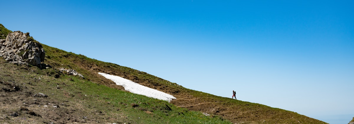 A lone hiker walks along a grassy mountain slope with a patch of snow under a clear blue sky. One side of the mountain features rocky terrain, while the other is lush with green grass. The landscape is expansive and serene.