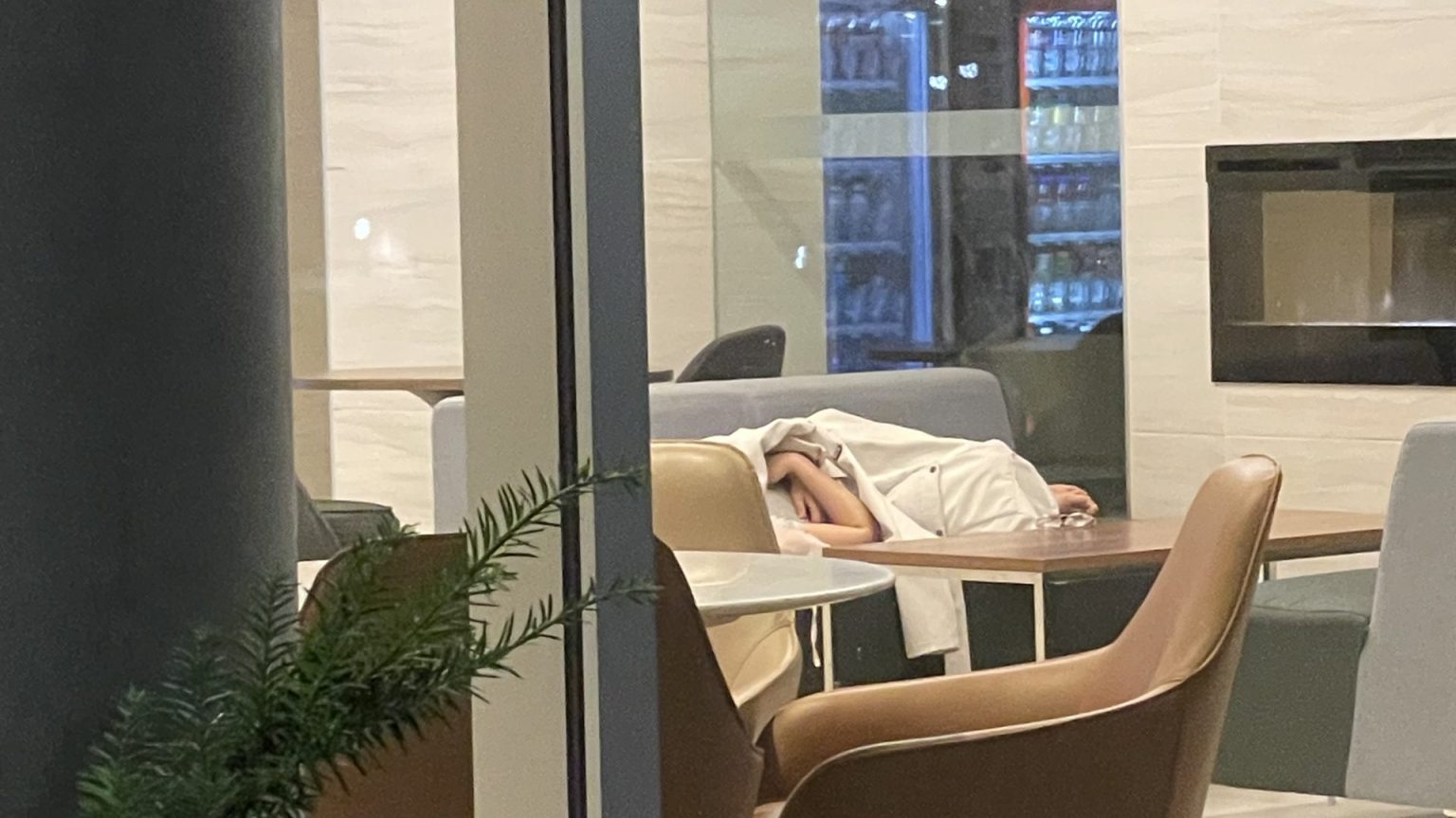 Person sleeping on a couch in a lounge area with a blanket covering them, likely escaping the heat wave outside. Adjacent is a table with a chair and a plant in the foreground. In the background, there is a wall with a large screen and a glass window revealing vending machines.