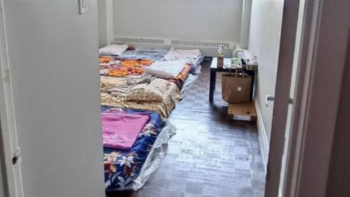 A room with several pillows and blankets arranged on the floor in a makeshift sleeping area, reminiscent of student living at the University of Waterloo. There is a small table at the far end of the room with various items on top and a cardboard box beside it. The wooden floor adds to the sparsely furnished housing landscape.