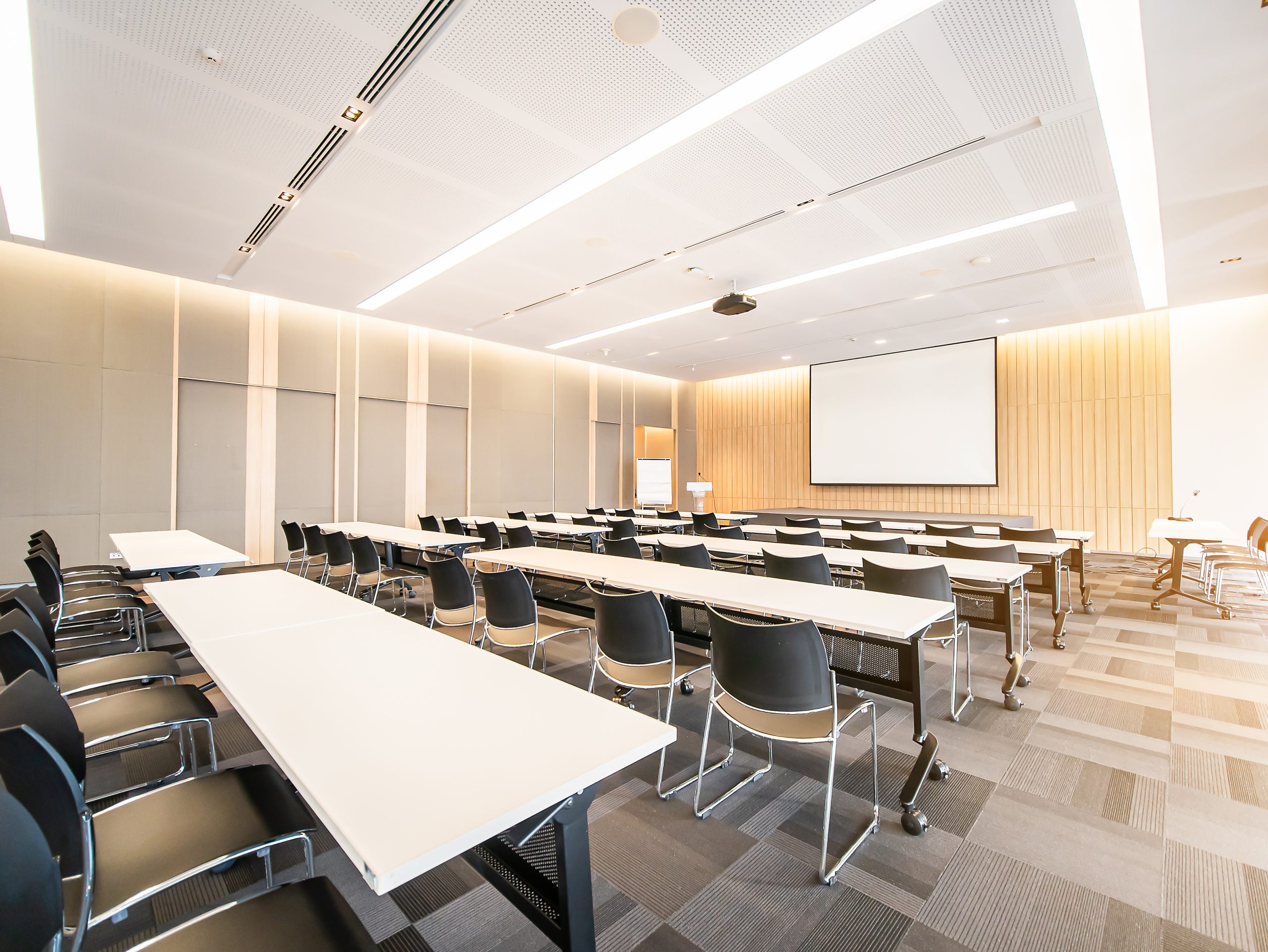 A modern, well-lit conference room with rows of black chairs and white tables arranged classroom-style facing a large projection screen awaits the July board meeting. The room has a neutral color palette with light wood paneling and a carpeted floor. Ceiling lights provide ample illumination.