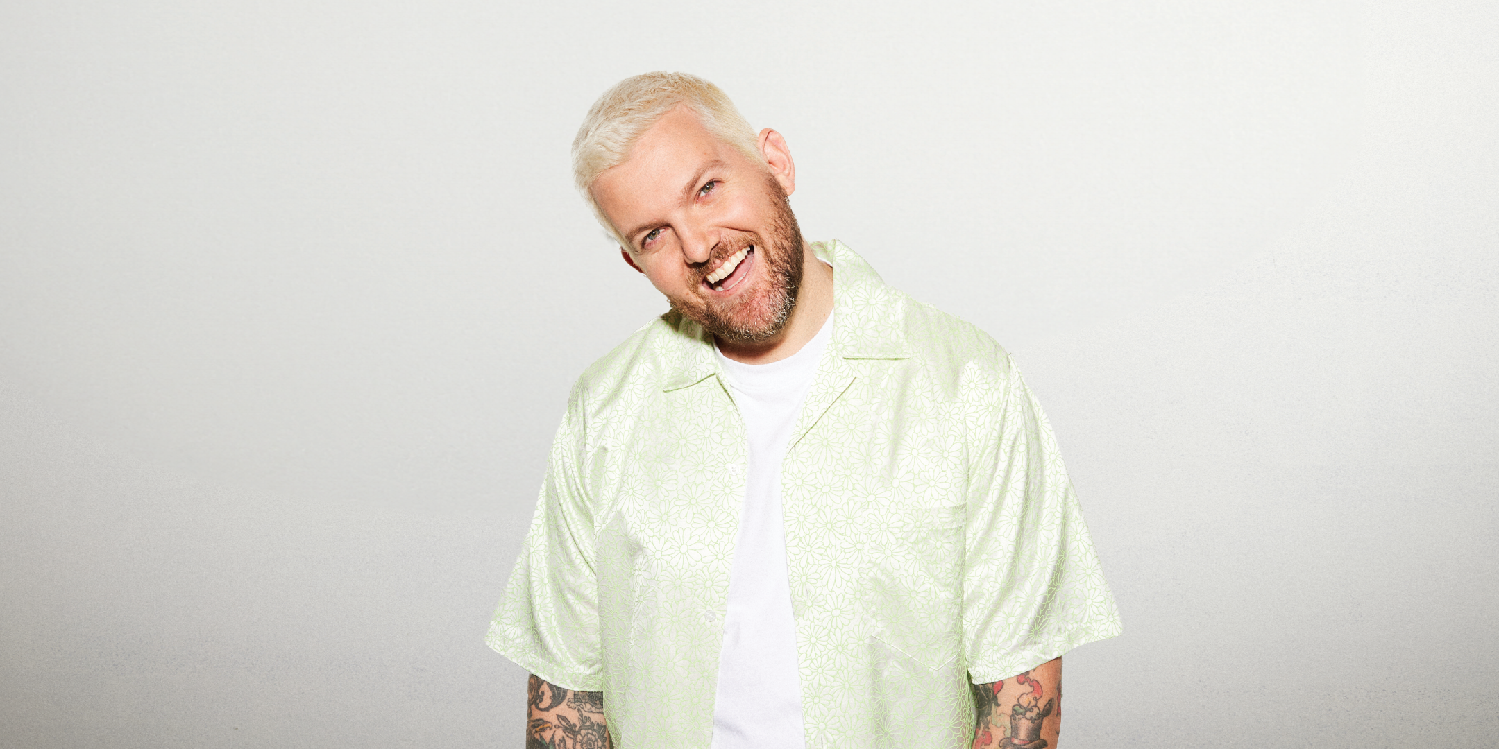 A bearded man with short platinum blonde hair smiles and slightly tilts his head to the side. He is wearing a light green short-sleeved button-up shirt over a white T-shirt. His arms, adorned with tattoos, are visible. The background is plain light gray.