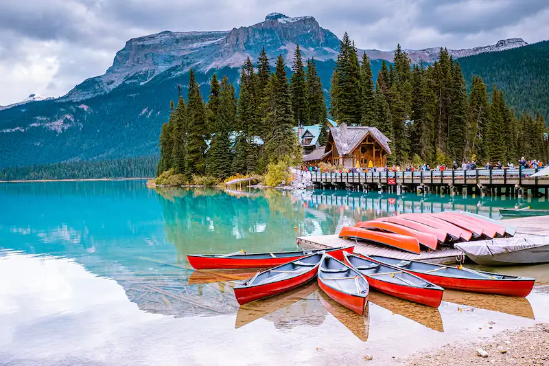 A serene lake surrounded by a forest and mountains, with several red canoes on the shore. A wooden cabin and dock are nestled among the trees, and the turquoise water reflects the cloudy sky and surrounding landscape.