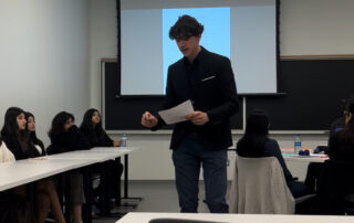 A person stands presenting in front of a group in a classroom setting, preparing for the Black & Gold Invitational's Mock Trial competition.