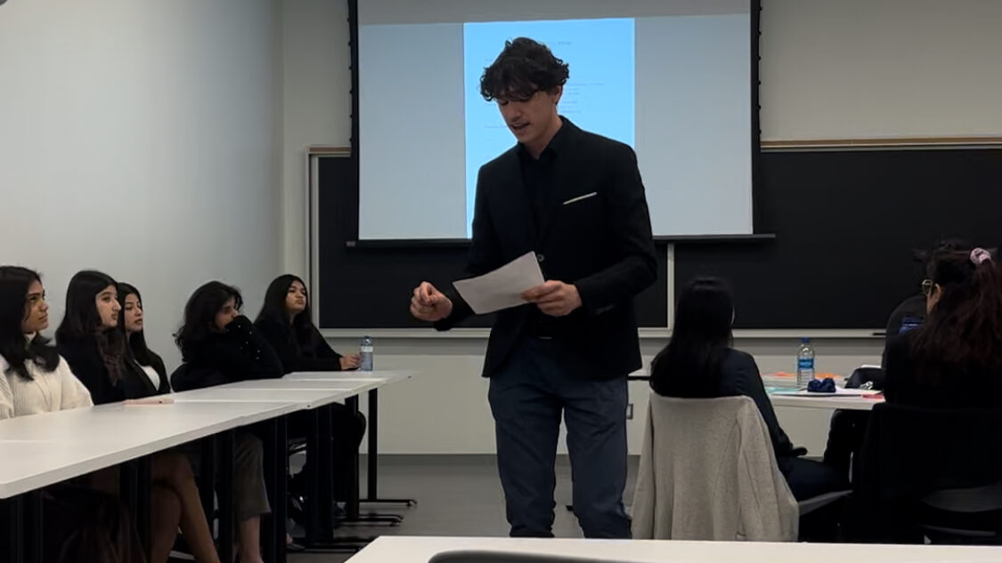 A person stands presenting in front of a group in a classroom setting, preparing for the Black & Gold Invitational's Mock Trial competition.