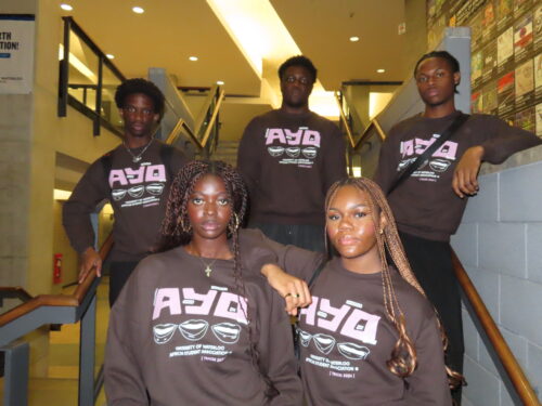 Five individuals wearing matching TRACES 2024 "Ayo" Sweaters stand together on a staircase, three on the steps and two at the bottom, posing for a group photo. Their stylish sweaters make them look perfectly coordinated.