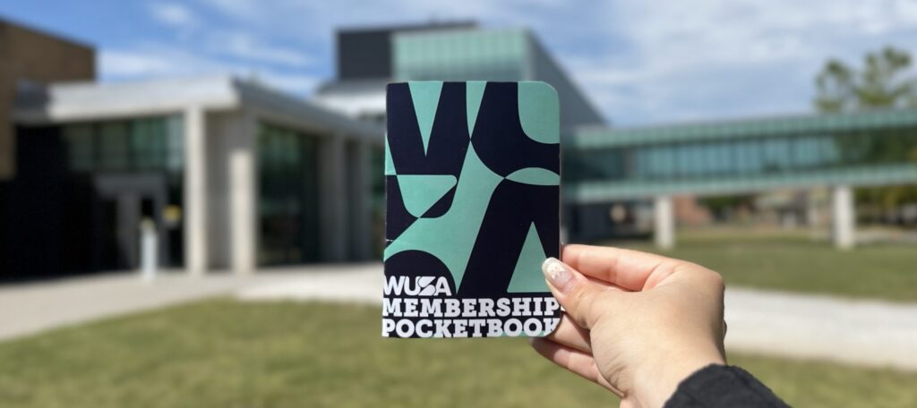 A person proudly holds a WUSA Membership Pocket Book in front of a modern building on a sunny day.