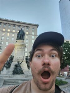 A very surprised President Nick with a hat on and pointing at a historic statue behind him.