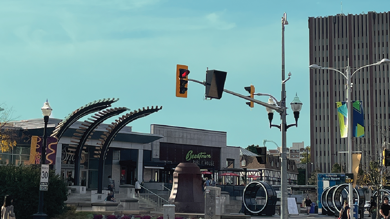 A vibrant city street scene in Uptown Waterloo, featuring modern buildings, streetlights, and a statue against a bright blue sky with traffic lights completing the urban panorama.