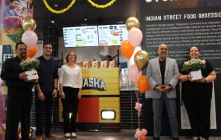 Five WUSA staff stand in the newly opened Chaska restaurant in SLC, holding gifts and flowers. Balloons add to the festive atmosphere, while the Chaska storefront and a menu board serves as a backdrop for this celebratory gathering.