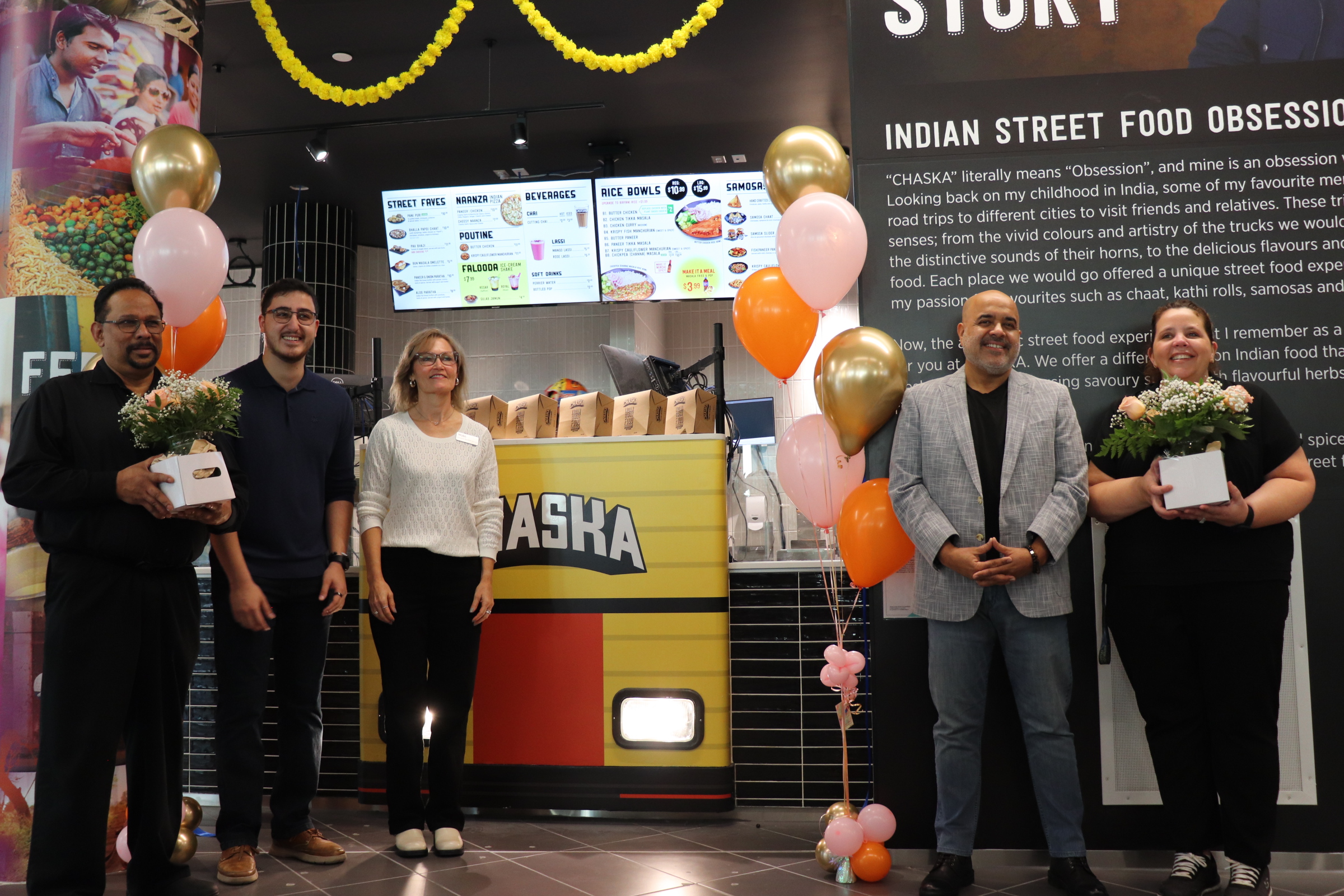 Five WUSA staff stand in the newly opened Chaska restaurant in SLC, holding gifts and flowers. Balloons add to the festive atmosphere, while the Chaska storefront and a menu board serves as a backdrop for this celebratory gathering.