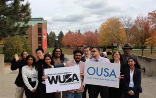 Amidst the vibrant autumn backdrop of colorful trees, a group of students proudly hold signs for WUSA and OUSA, highlighting their spirited enthusiasm.