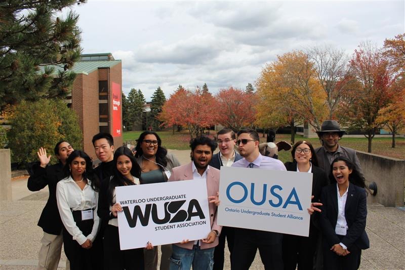 Amidst the vibrant autumn backdrop of colorful trees, a group of students proudly hold signs for WUSA and OUSA, highlighting their spirited enthusiasm.