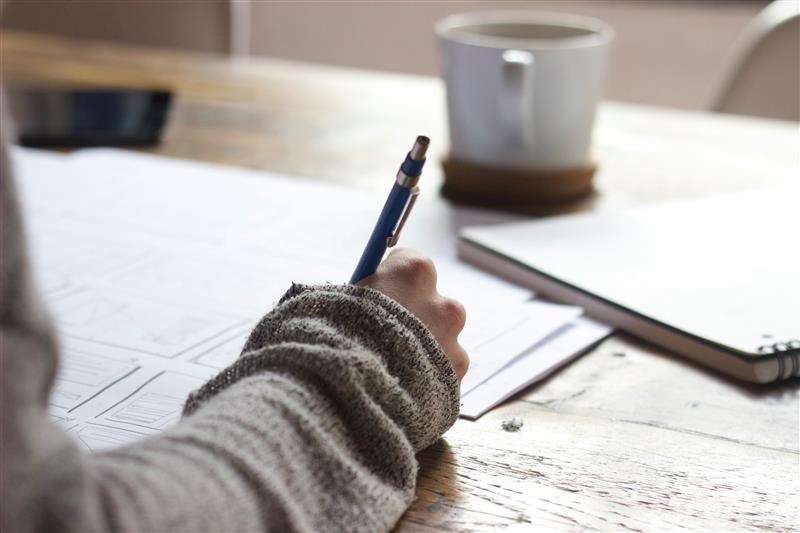 A student is diligently writing on a paper at a wooden desk, with a steaming cup and an open notebook nearby.