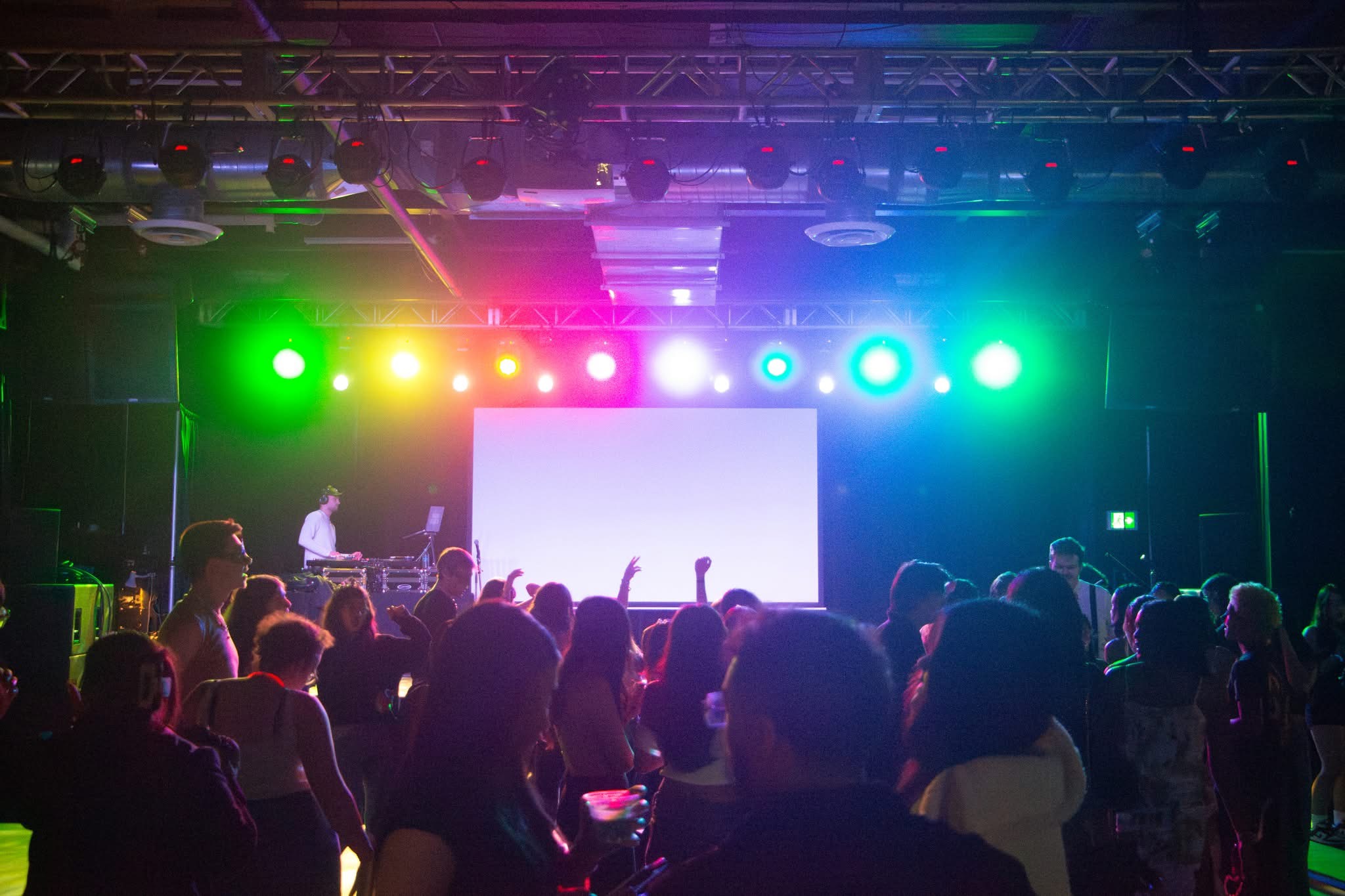 People dancing in a dimly lit club with colorful stage lights and a DJ, showcasing the latest fashion trends on the vibrant dance floor.